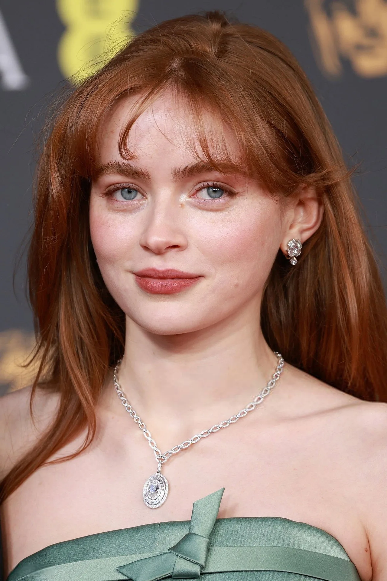 Sadie Sink vestindo roupa de treino preta, segurando halteres, ao fundo pôster de Spider‑Man e luzes vermelhas