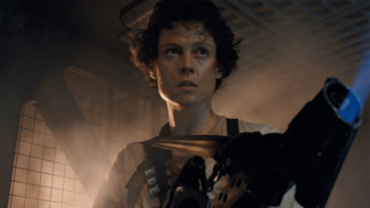 Mulher levantando peso com camiseta de alienígenas, ao fundo poster de “Aliens” e barra de supino