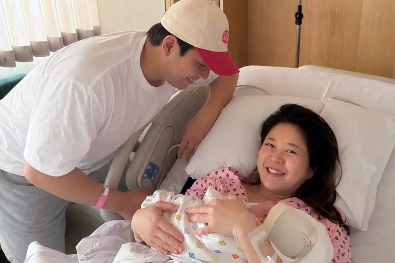 Kim Seung Hye abraça a filha recém‑nascida enquanto Kim Hae Jun sorri ao seu lado, ambos em roupa casual