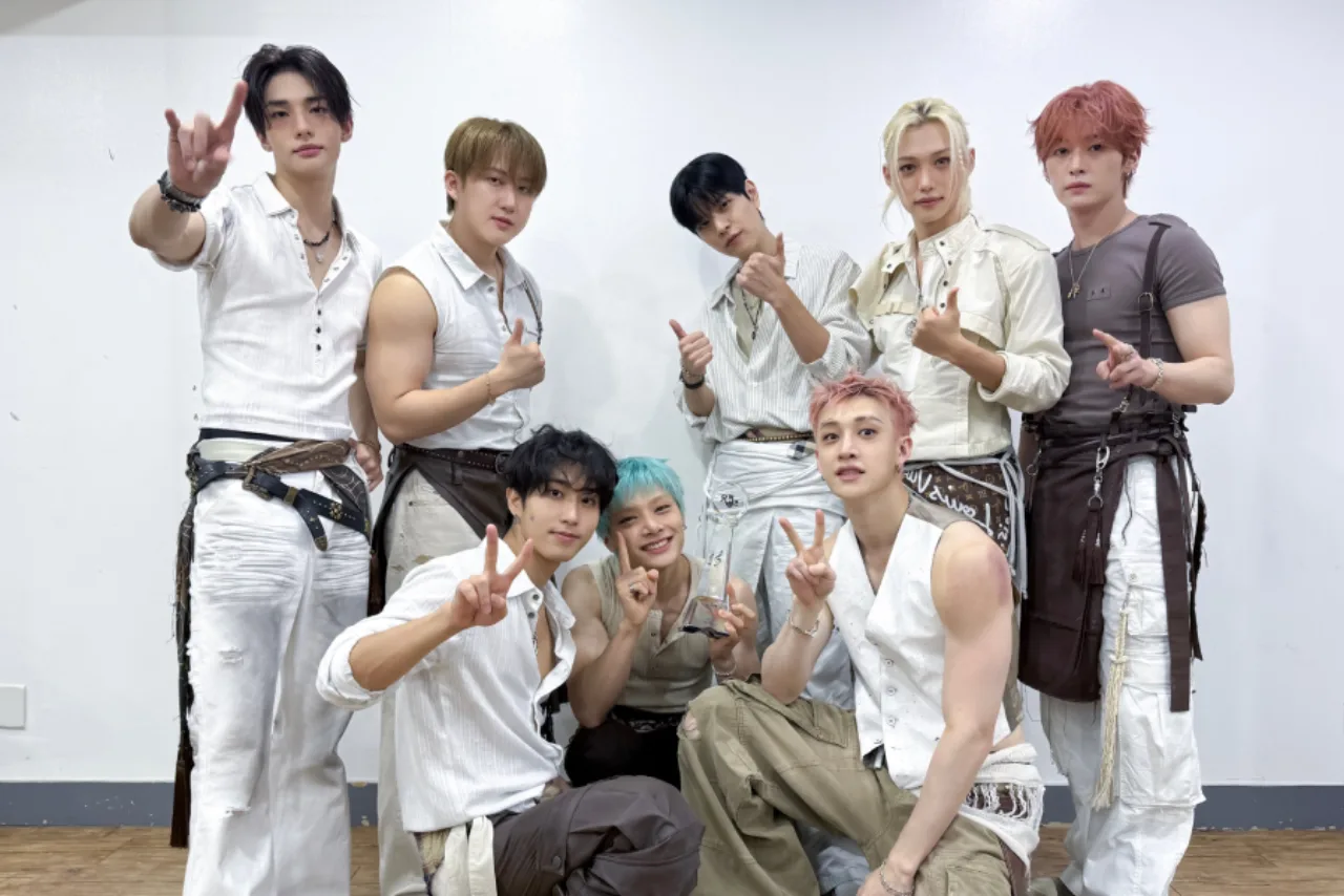 Jovem usando camiseta do Stray Kids faz agachamento com halteres, fones de ouvido e garrafa de água ao fundo