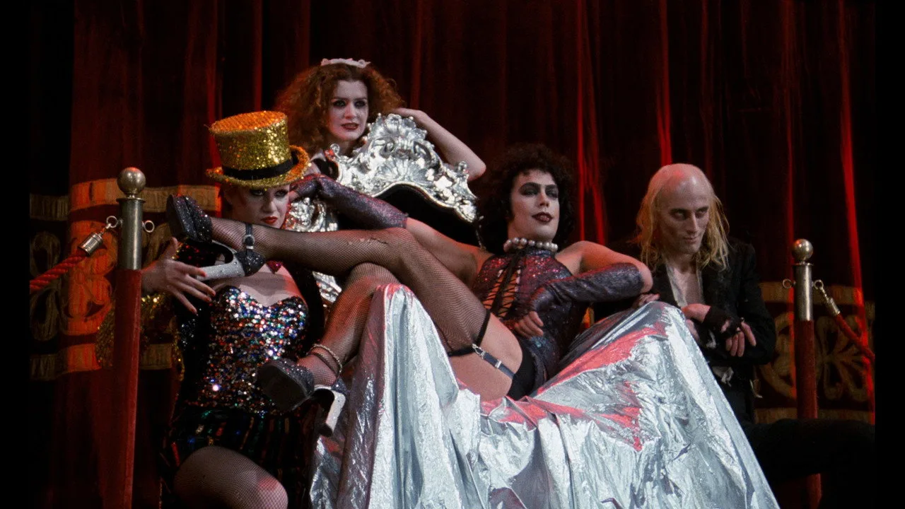 Jovem fazendo prancha ao lado de um prato com salada colorida e garrafa d'água, enquanto tela exibe Tim Curry
