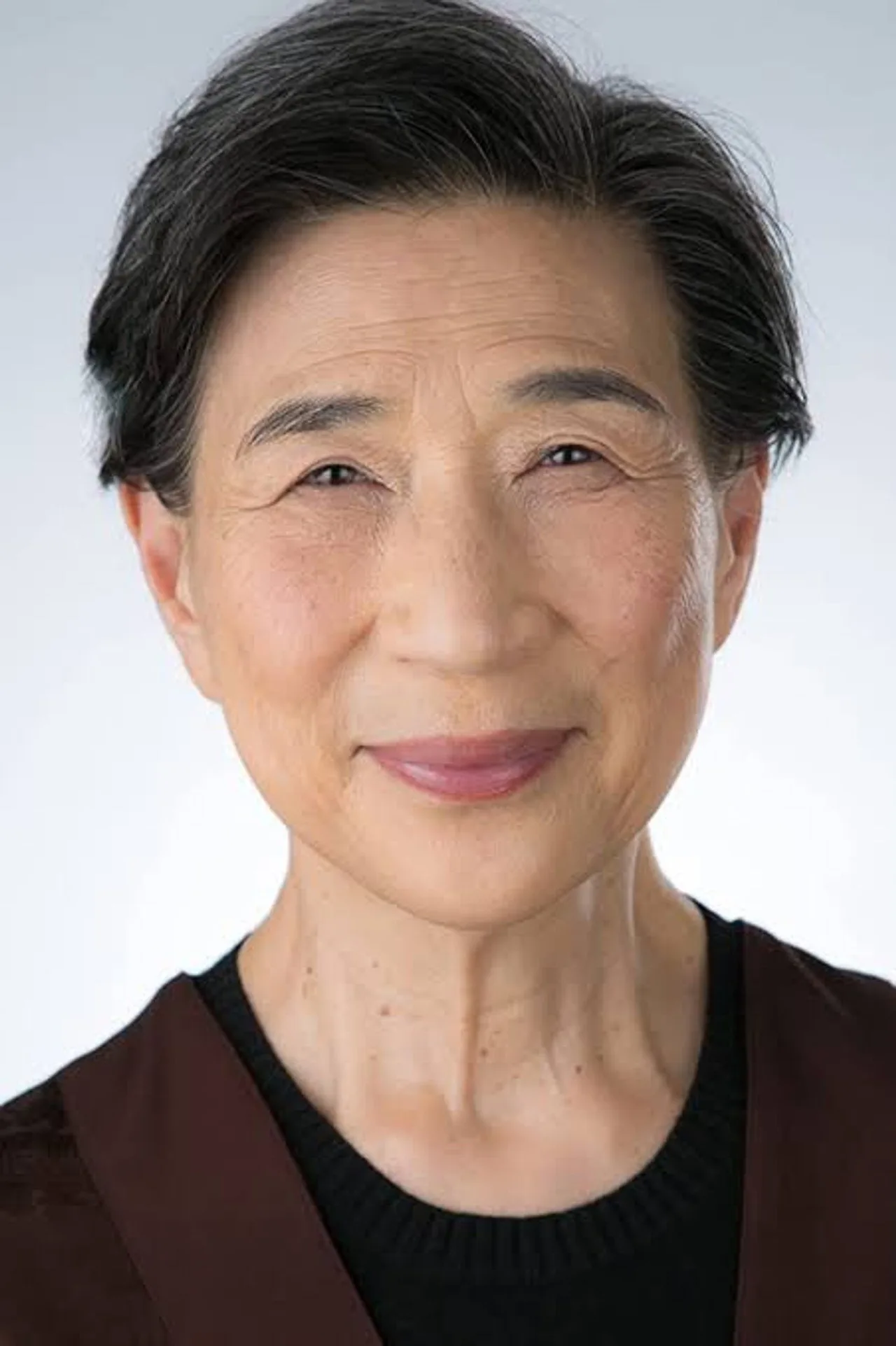 Wai Ching Ho em close-up, cabelos grisalhos ao vento, vestindo jaqueta de couro sobre camisa branca, segurando um script