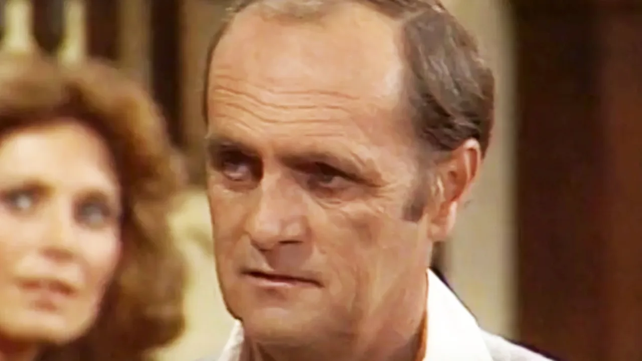 Ator idoso praticando yoga em casa, vestindo camiseta com logo da série Newhart