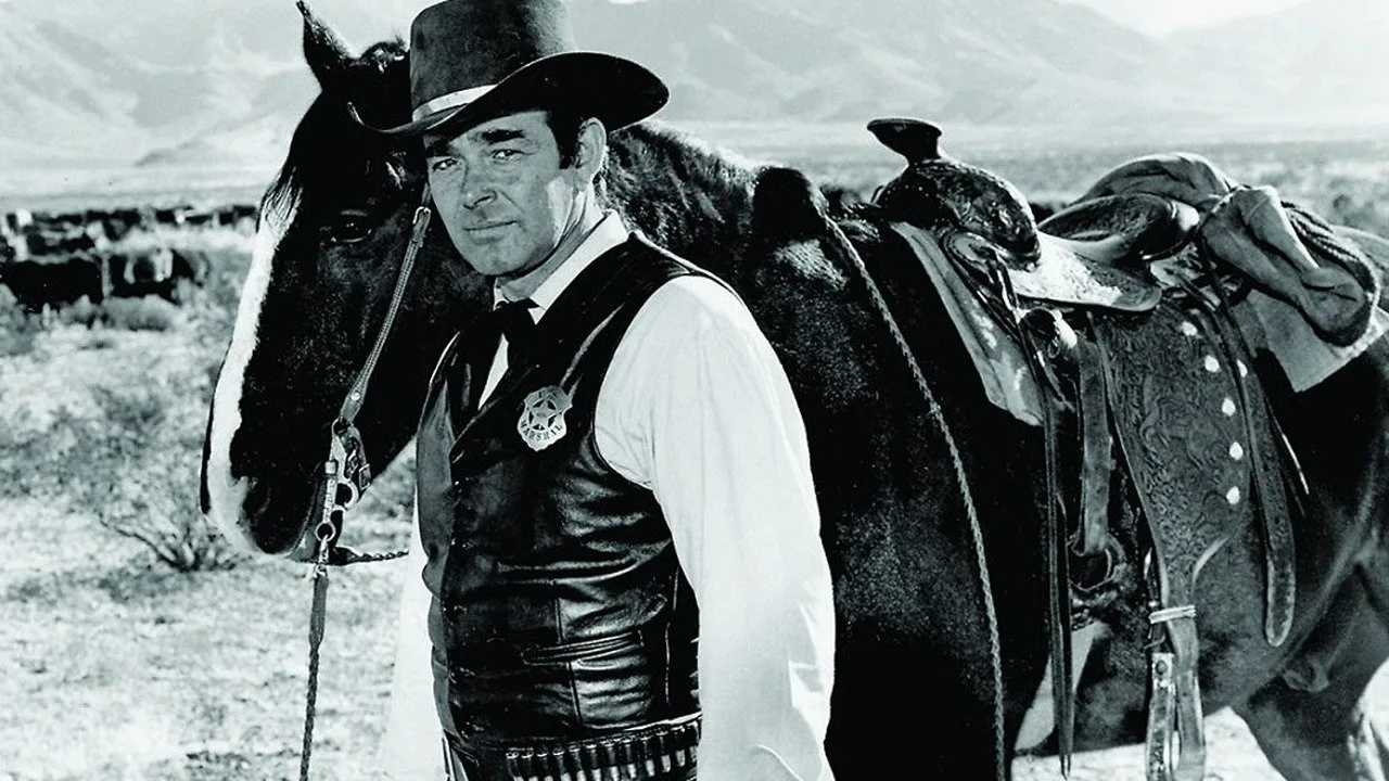 Homem musculoso vestindo chapéu de cowboy faz agachamento com corda de laço ao pôr‑sol no deserto