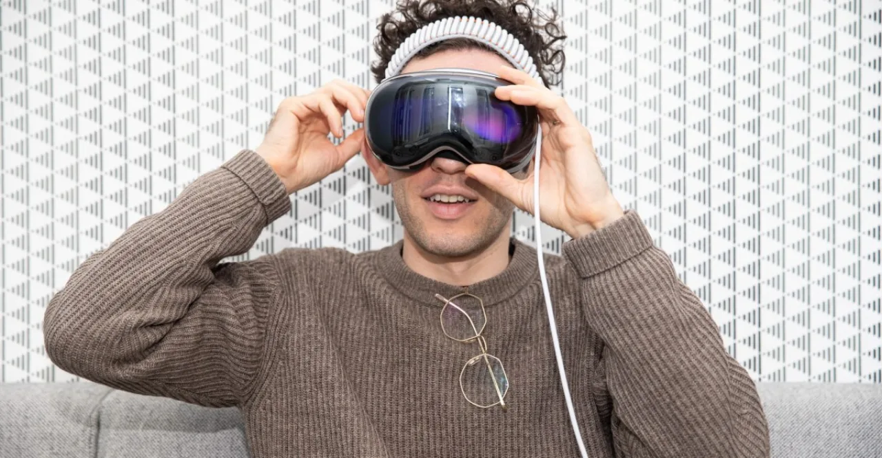Jovem fazendo alongamento com óculos Vision Pro, laptop Apple aberto ao fundo e garrafa de água ao lado
