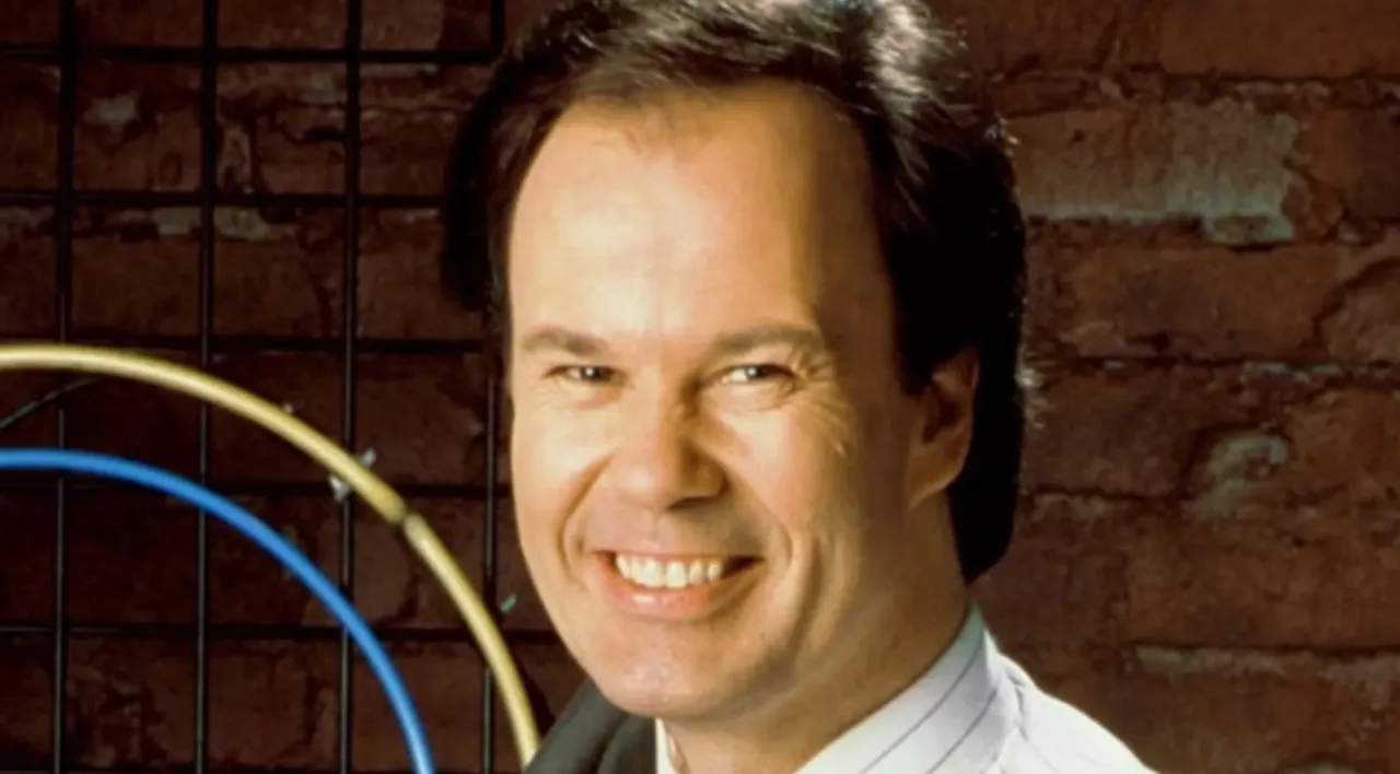 Dennis Haskins, em traje azul, posando como Sr. Belding, com o logo de “Saved by the Bell” ao fundo