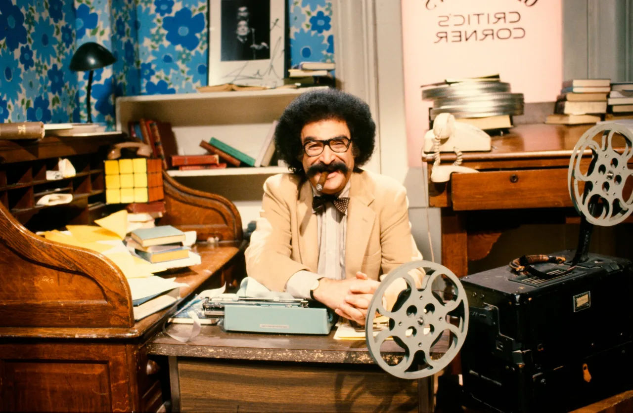 Gene Shalit sorrindo, usando óculos, segurando uma maçã verde ao lado de halteres