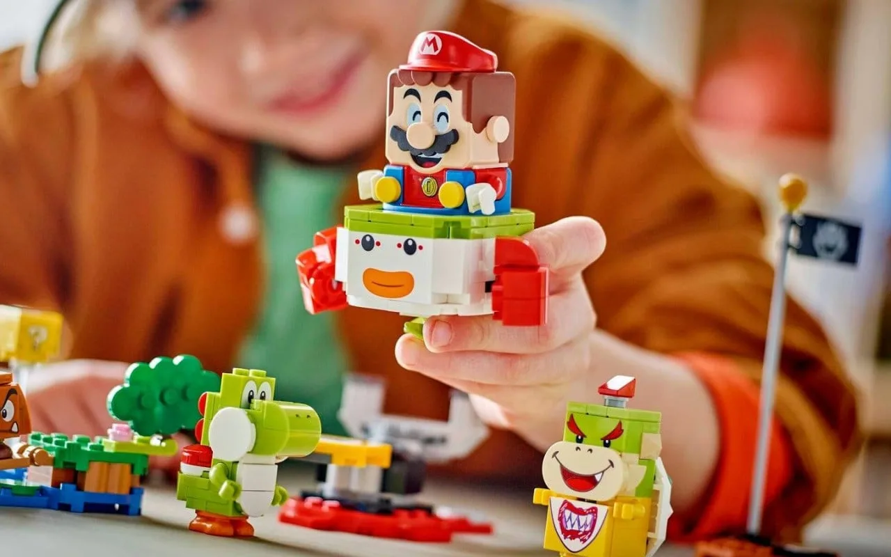 Pessoa montando um set LEGO do Mario sobre uma mesa organizada ao lado de peças coloridas e um controle de videogame