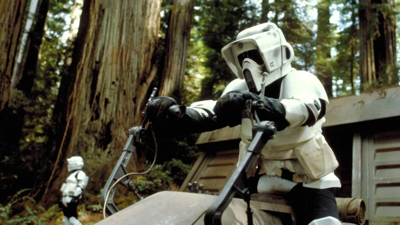 Jovem em bicicleta estática, vestindo roupa de ciclismo, segura a pistola laser Kenner Biker Scout ao lado da caixa transparente