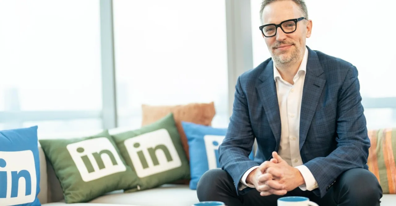 Ryan Roslansky em pé, ao lado dos logotipos da Microsoft, Office e Teams, em traje executivo