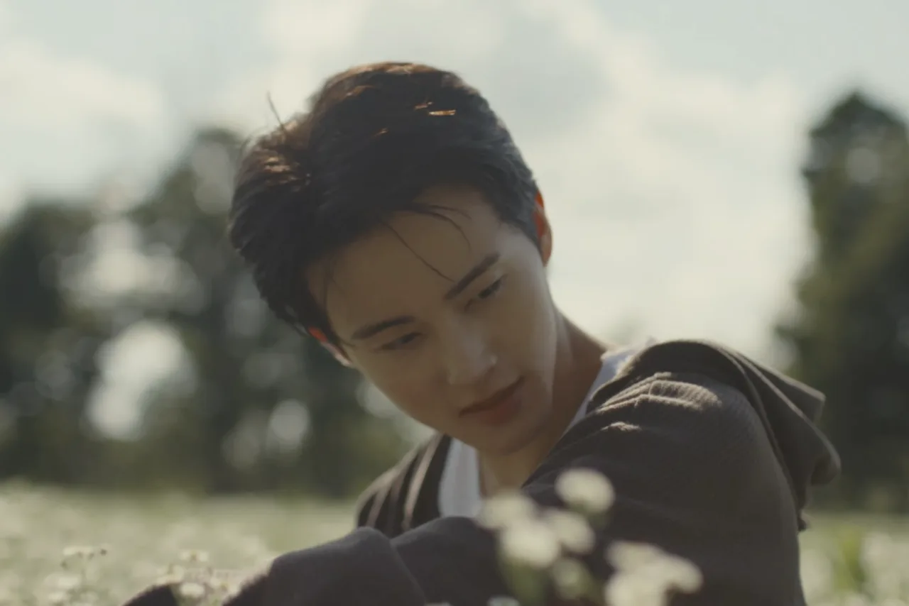 Jovem com fones, fazendo agachamento com halteres, garrafa d'água e laptop exibindo o teaser de Mark Lee