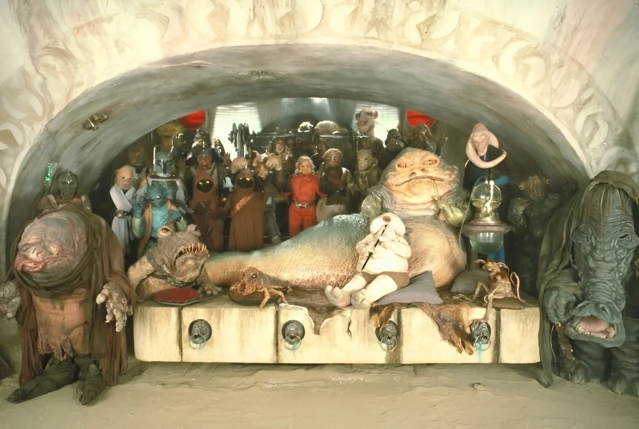 Homem fazendo supino com barra, ao fundo a caixa de brinquedo Jabba’s Dungeon aberta, revelando figuras colecionáveis