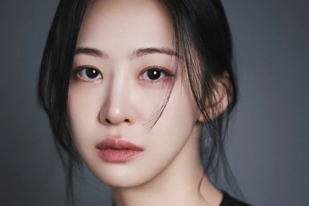 Dasom posa sorrindo em foto de perfil, vestindo roupa elegante, ao fundo o logotipo da The J’s E&M