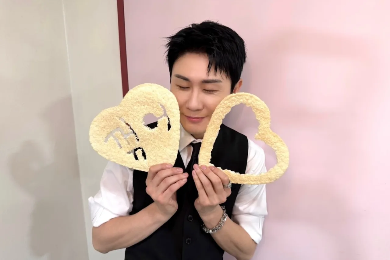 Young Tak ergue o troféu do Music Bank, ao fundo o logo do programa e luzes de palco vibrantes