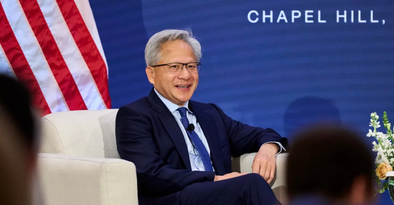 Jensen Huang no palco do All‑In Summit, segurando microfone enquanto a tela exibe a foto de Donald Trump