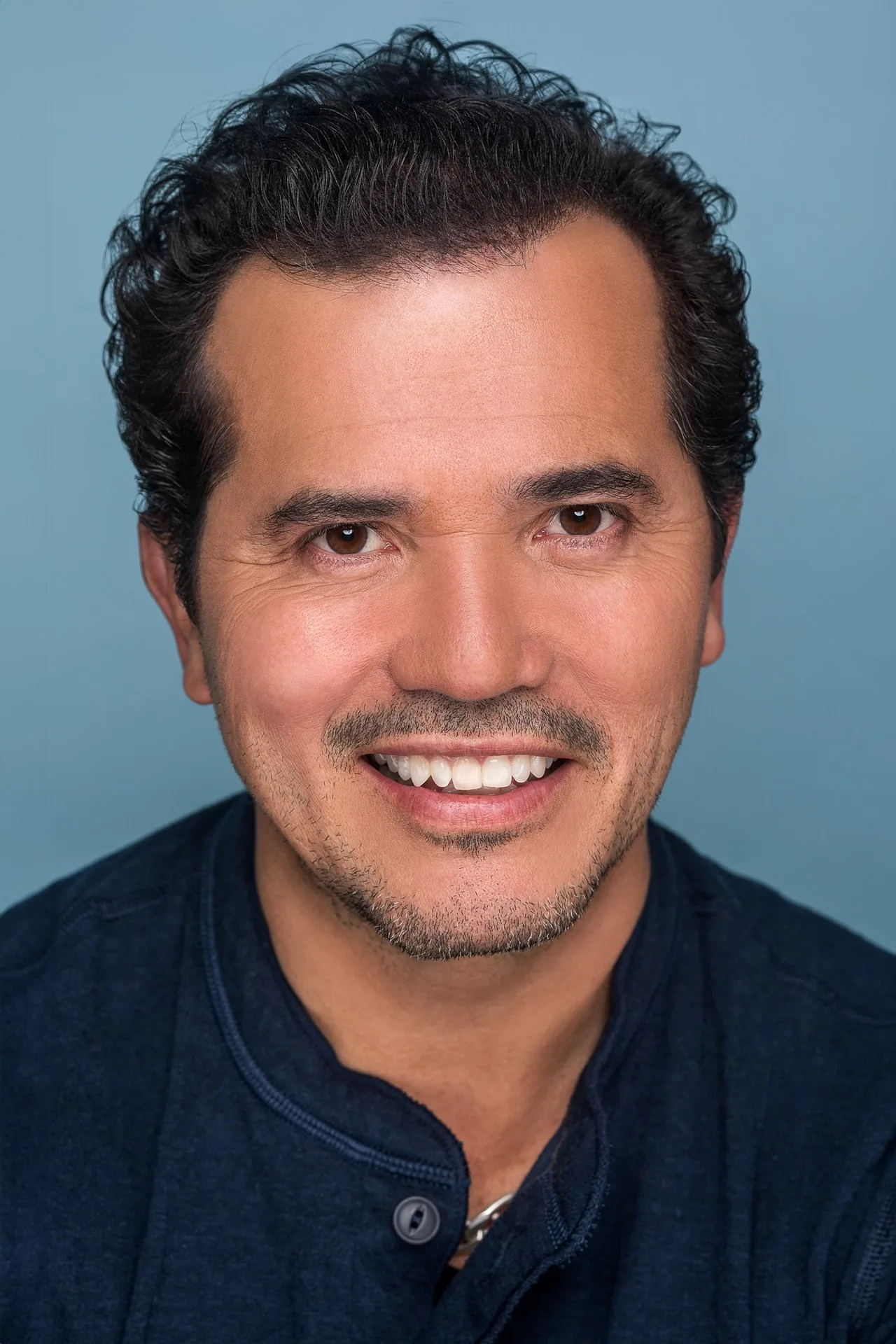 John Leguizamo em roupa esportiva, segurando halteres, ao lado de um tapete de yoga numa academia moderna