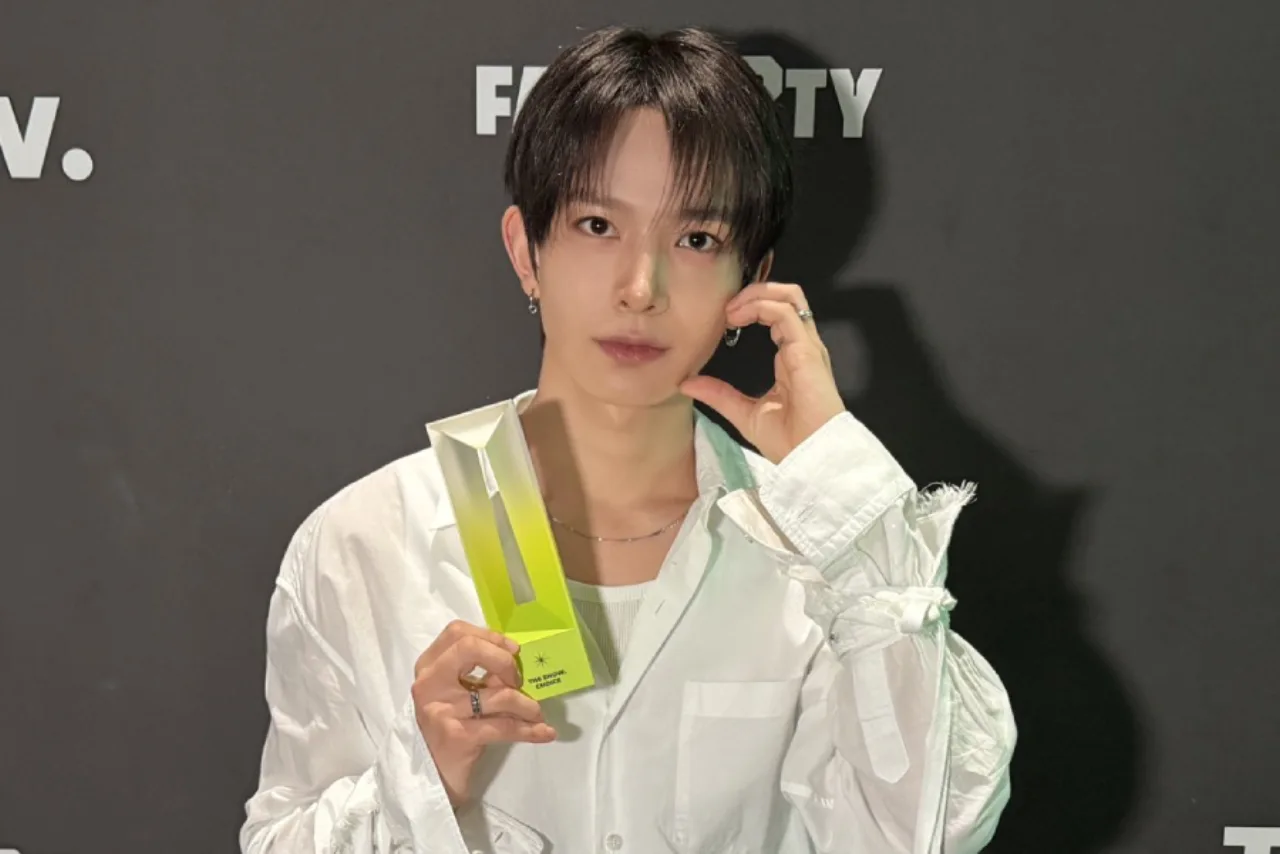 Heeseung ergue o troféu do The Show, sorrindo sob luzes neon, com o logo do programa ao fundo