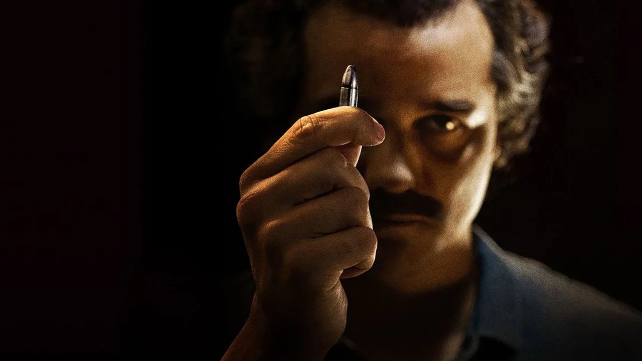 Um homem em traje de corrida, segurando um controle remoto, com pôsteres de Narcos e The Westies ao fundo
