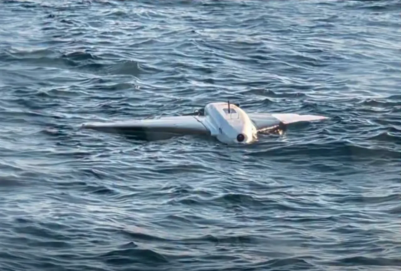 Um mergulhador em traje, segurando um drone desativado ao lado de equipamentos de mergulho e plataforma offshore