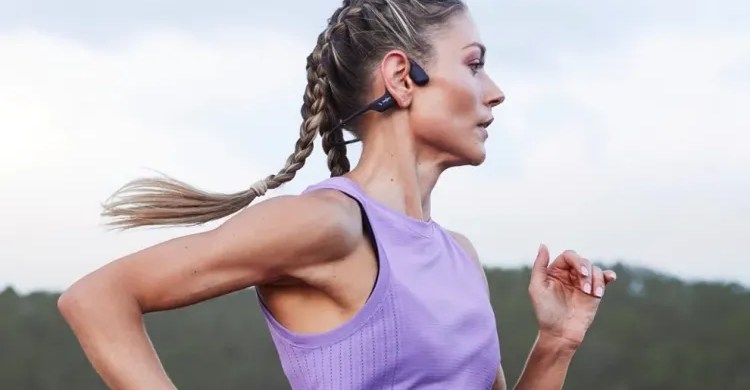 Fones Shokz OpenRun Pro presos ao crânio de corredor em pista, ao lado de garrafa d'água e smartwatch